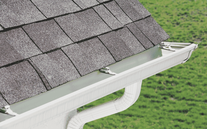 Alt Text: Beige residential house with asphalt shingle roof and white gutter system under a clear blue sky